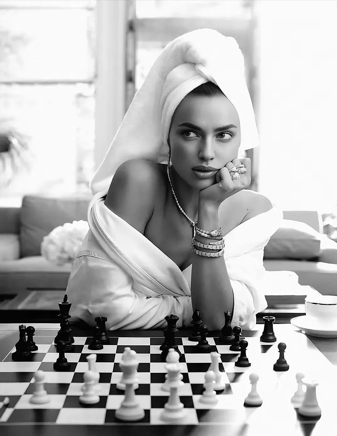 A woman in a white robe poses thoughtfully while playing a chessboard on a table.