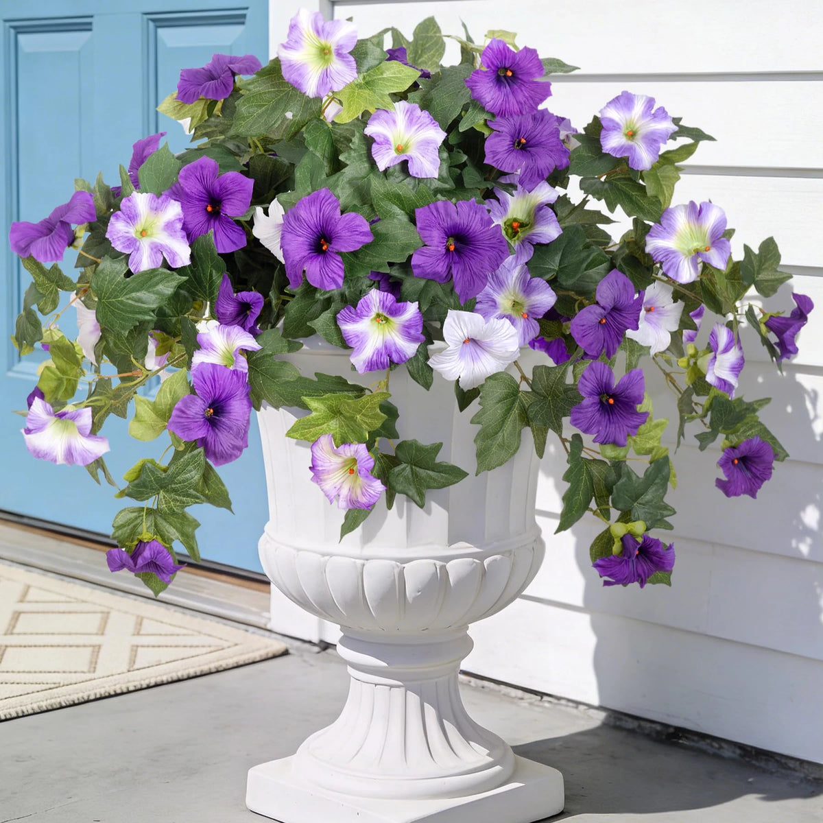 A white pedestal planter filled with purple and lavender artificial petunia bundles outdoors.