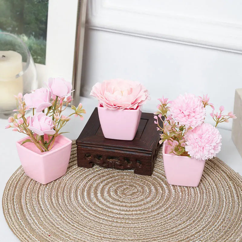 Three pink faux bonsai plants in small pots displayed on a woven mat.