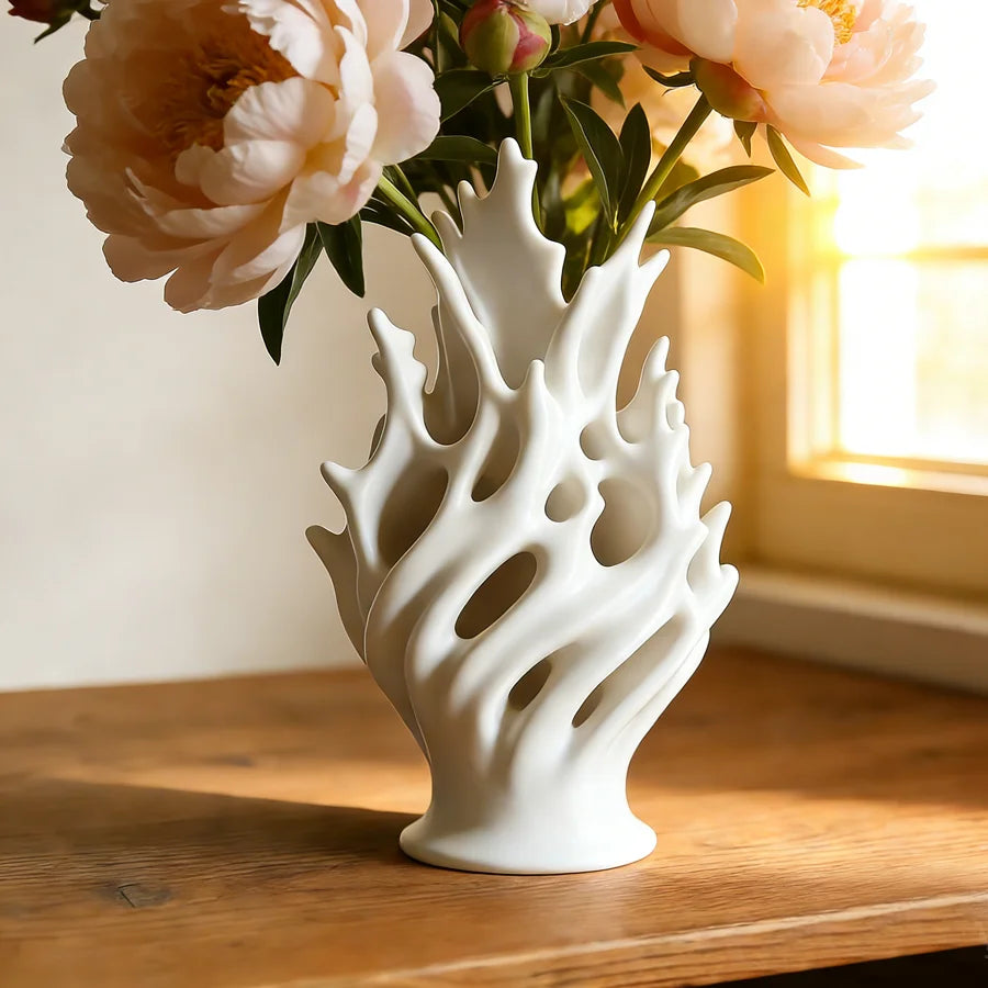 A white, sculptural plastic flower vase holding soft pink peonies on a wooden table near a window.