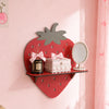 A red strawberry shaped wooden wall shelf featuring a mirror and pink floral accents on a light pink wall.