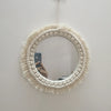 Miroir rond encadré par une corde macramé crème épaisse et des franges, accroché à un mur blanc.
