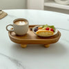 A white ceramic mug filled with latte art sits on a wooden tray next to a small plate of fruit and greens.