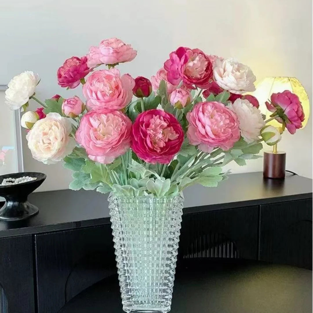 A bouquet of pink and white silk peony flowers in a clear glass vase on a dark table.