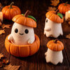 Three small white elf-like figures with orange pumpkin heads are displayed on a dark wooden surface surrounded by autumn leav