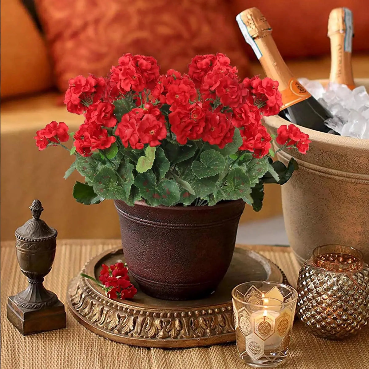 A potted silk begonia artificial flower with red blooms sits on a wooden tray next to a bottle and decorative glass.