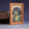 A framed watercolor painting of a green frog in a wooden frame displayed on a dark surface.
