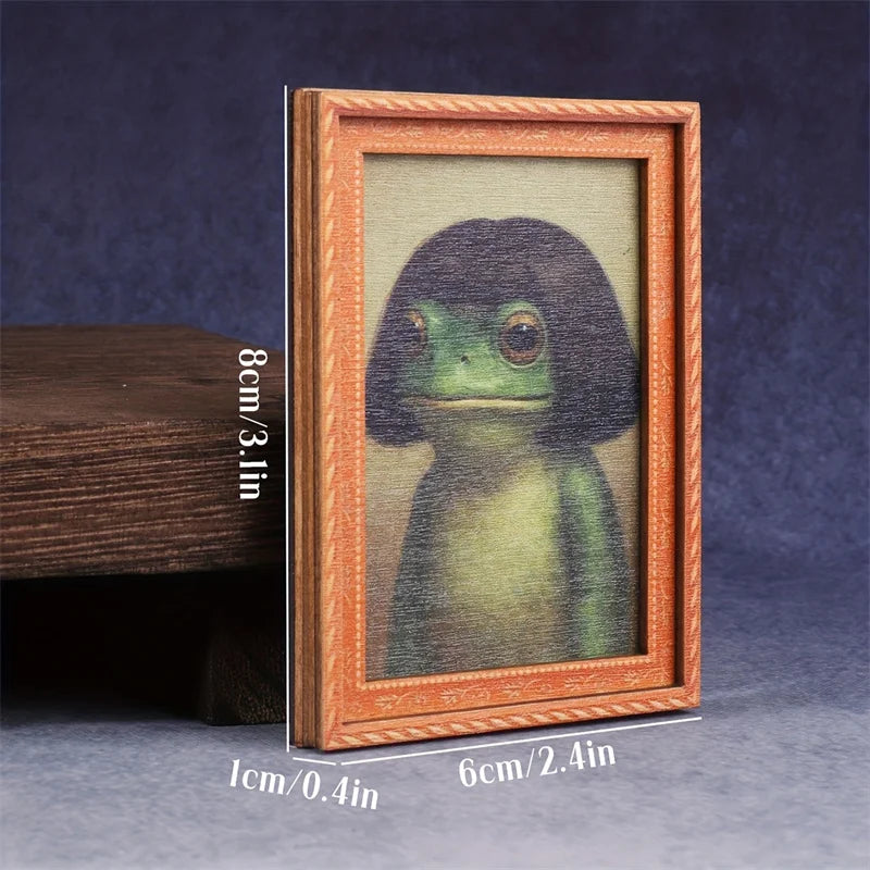 A framed watercolor painting of a green frog in a wooden frame displayed on a dark surface.
