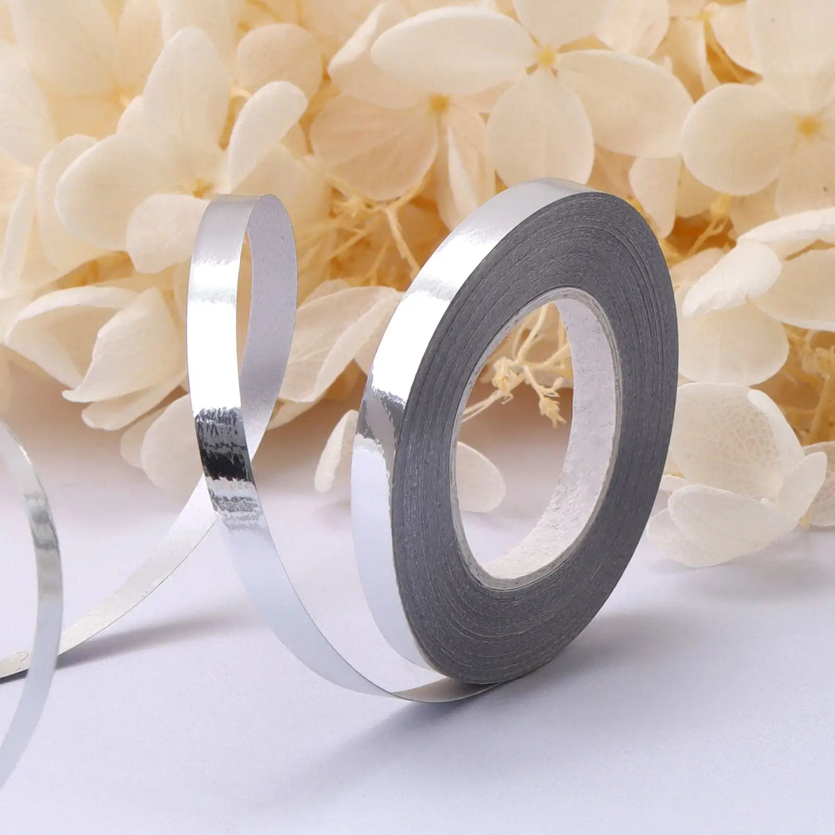 Roll of silver satin balloon ribbon lying on a white surface next to light yellow flowers