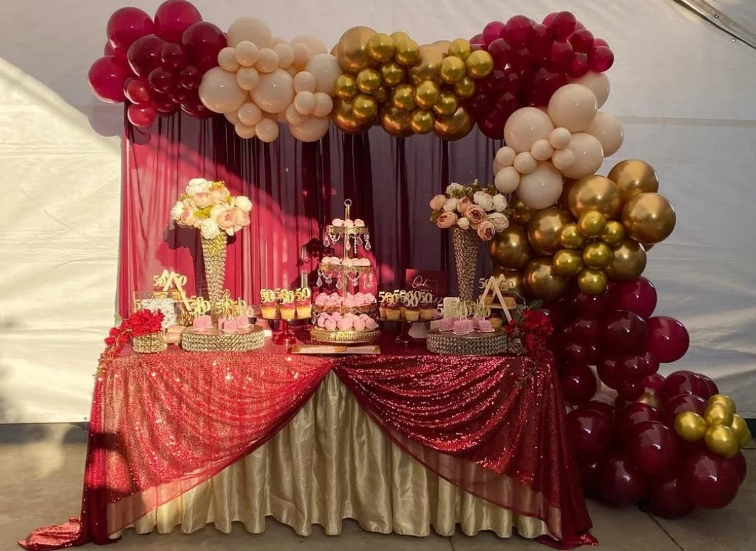 A balloon garland arch display featuring burgundy, gold, and white balloons on a draped table.