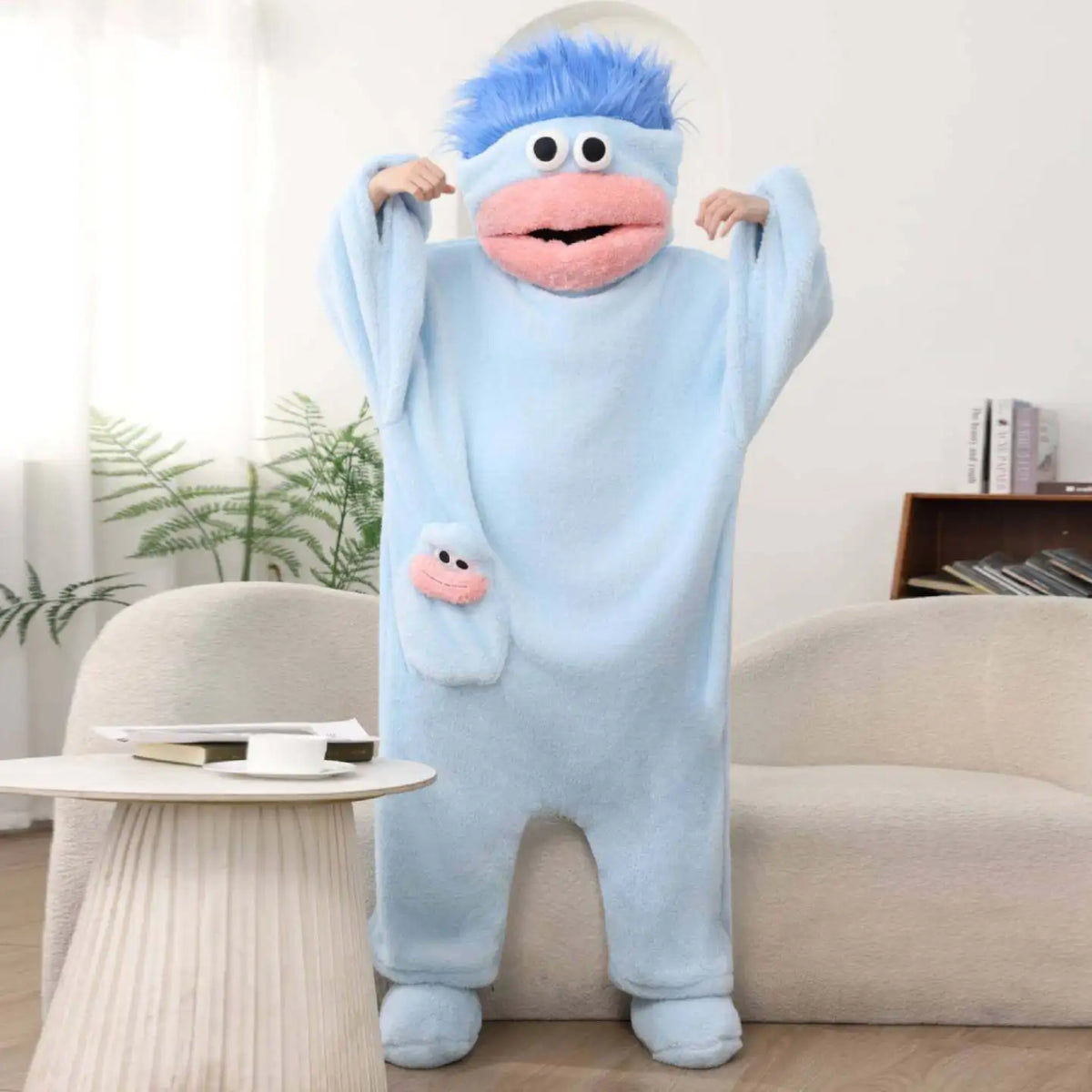 A light blue coral fleece hooded sleepsuit resembling a monster with pink features is displayed on a couch.