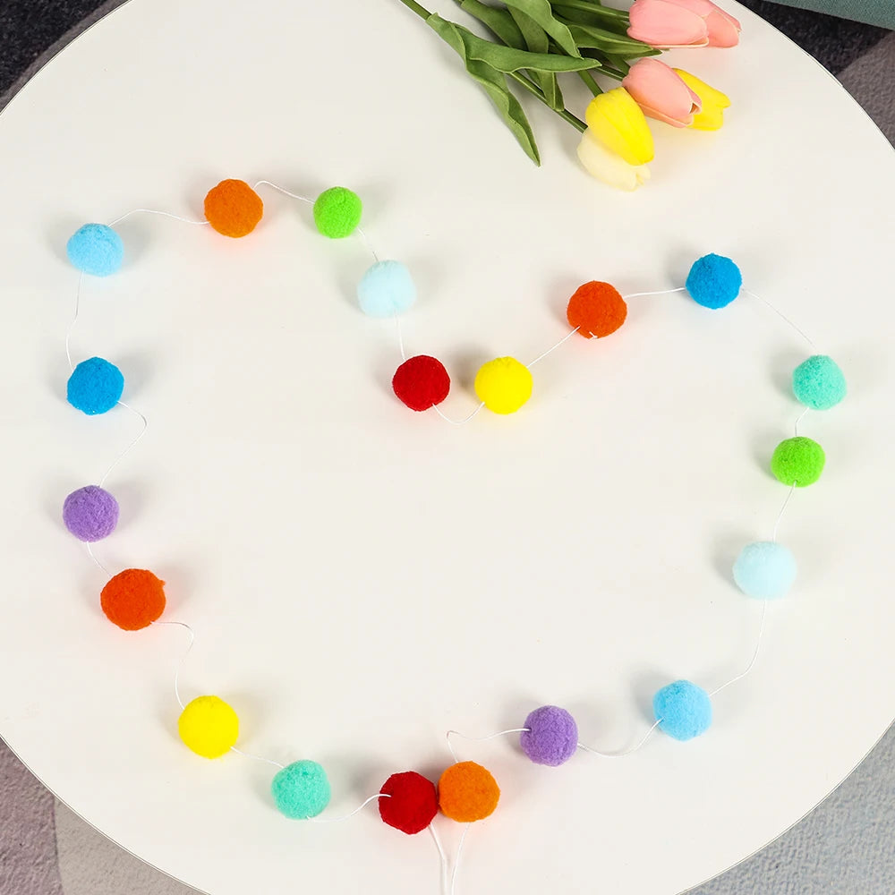 A heart-shaped garland made of colorful felt balls in shades of orange, yellow, green, and blue.