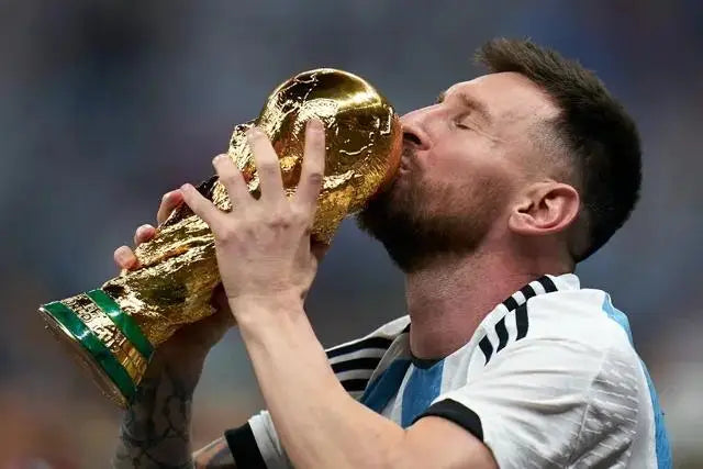 Leyenda del fútbol Lionel Messi sosteniendo un trofeo dorado con la boca, celebrando en el campo.