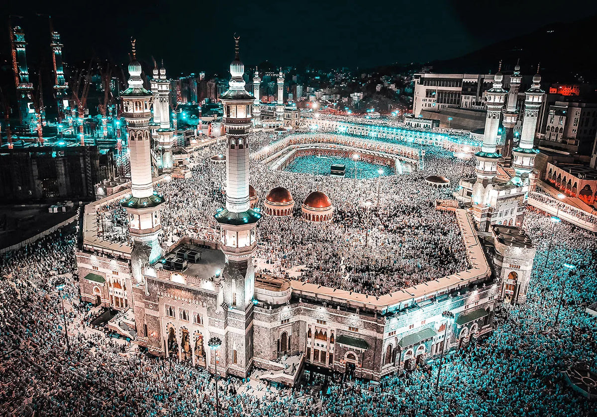 Night view of a mosque with minarets and a courtyard filled with people, illuminated by blue lights.