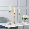 A set of three gold-toned, slender taper candles displayed on a white tabletop against a light gray wall.