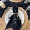 A stack of black fringed cotton napkins is displayed on a round woven mat with a white plate and a candle.