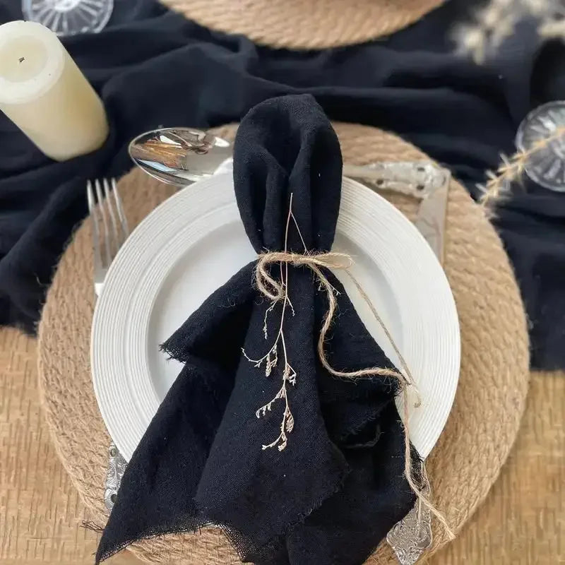 A stack of black fringed cotton napkins is displayed on a round woven mat with a white plate and a candle.