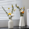 A white ceramic mini flower vase holds greenery and yellow flowers on a dark surface near a window.