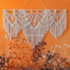 Handwoven boho macrame wall hanging tapestry in cream color displayed against an orange wall with blurred floral elements.