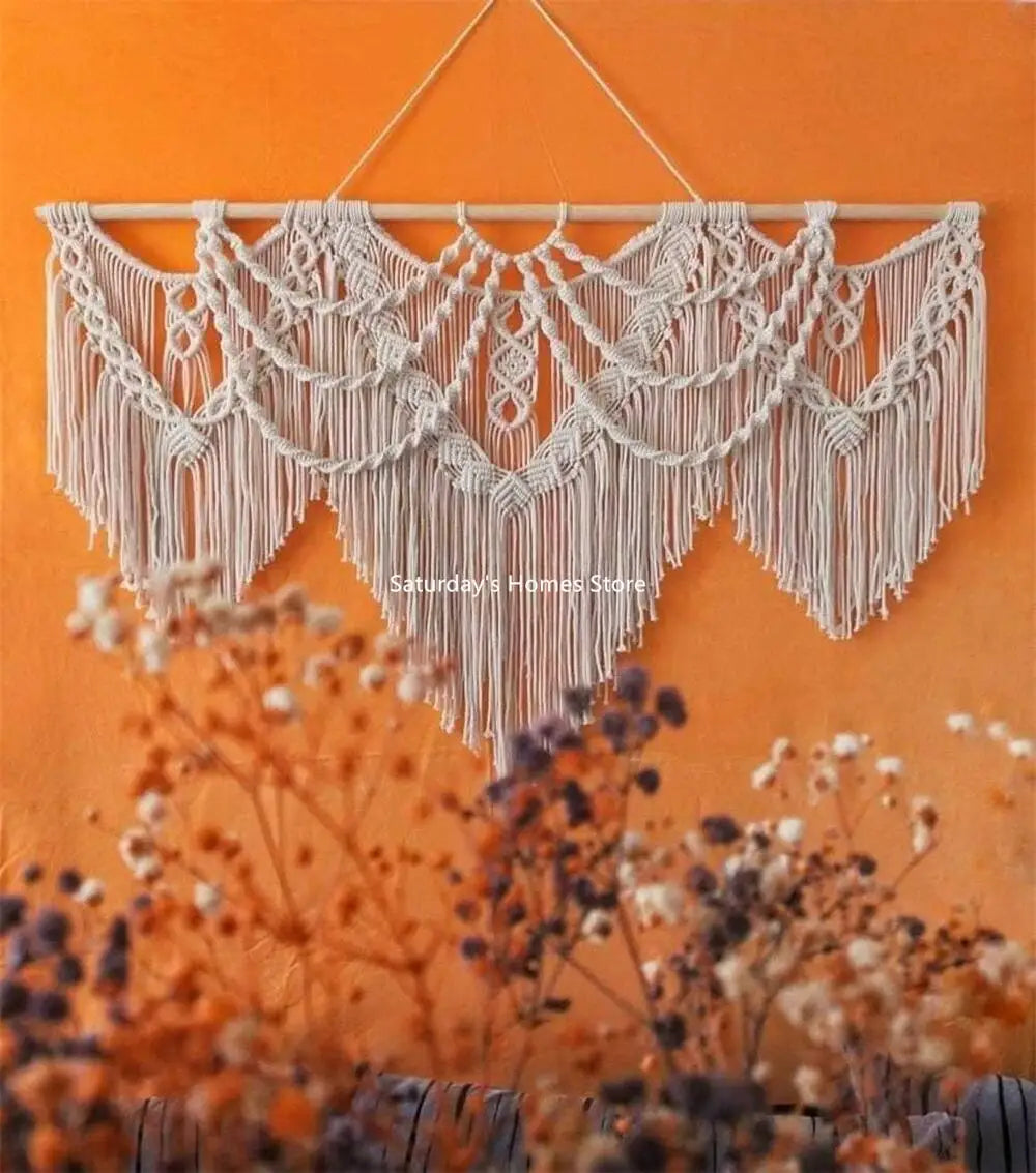 Handwoven boho macrame wall hanging tapestry in cream color displayed against an orange wall with blurred floral elements.