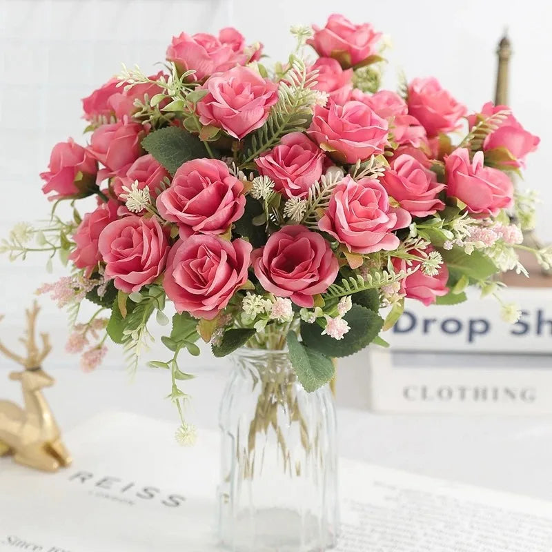 A bouquet of pink and coral silk artificial roses arranged in a clear glass vase on a white surface.