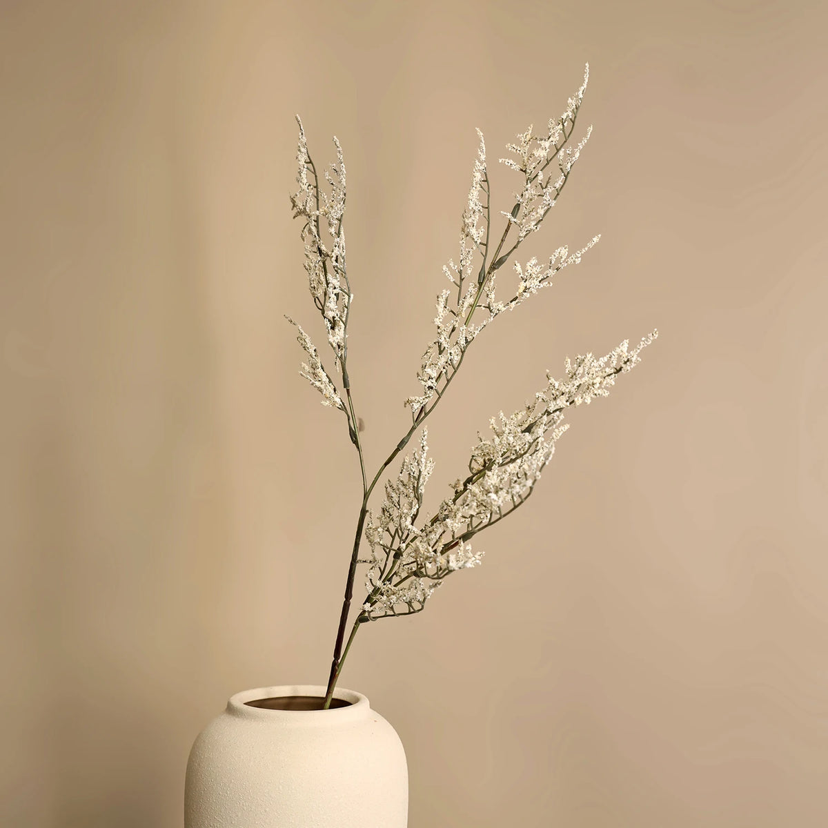 A sprig of artificial silk lover grass in a white ceramic vase against a neutral background.