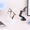 A minimalist black glass holder displaying a pair of round tortoiseshell eyeglasses on a white display stand.