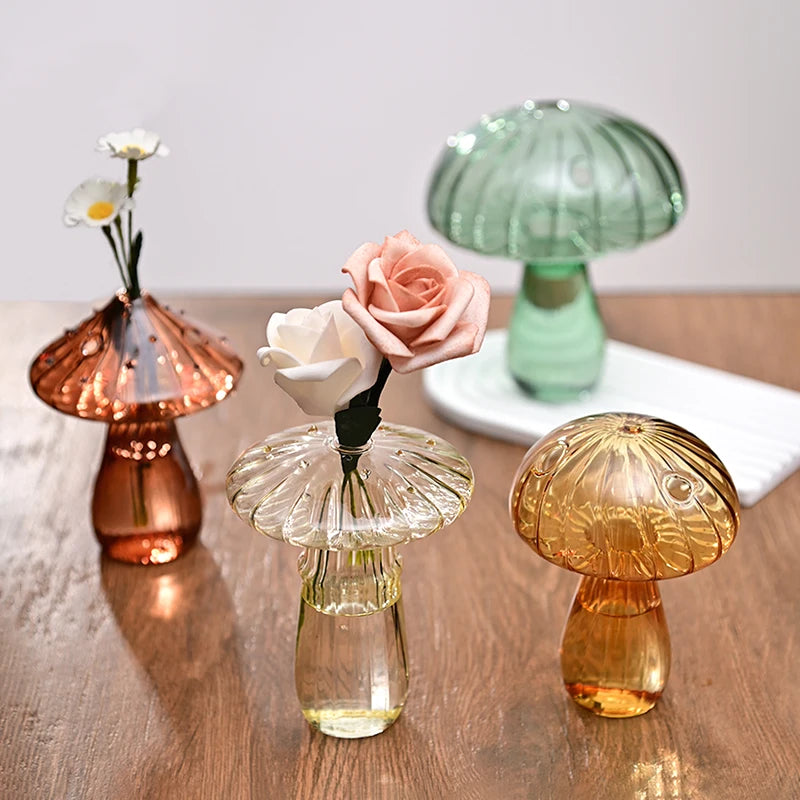 Trois vases en forme de champignons en verre, dans des tons rose gold, vert pâle et ambre, exposés sur une surface en bois.