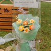 A handmade crochet flower bouquet featuring yellow and orange flowers wrapped in clear cellophane.