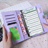 A purple leather binder with a budget sheet and cash envelope organizer is shown on a light pink surface.