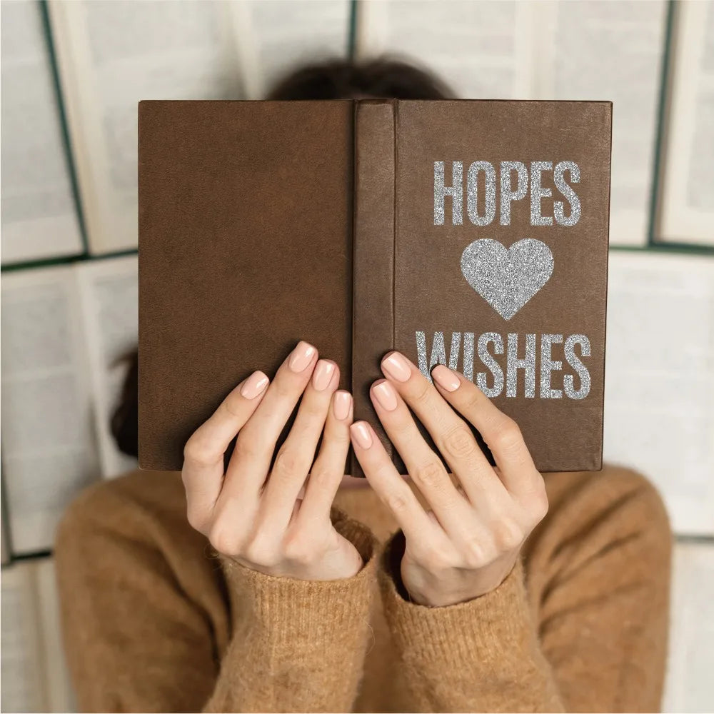 A person holds open a brown notebook displaying the words HOPES and WISHES in silver glitter alphabet letter stickers.