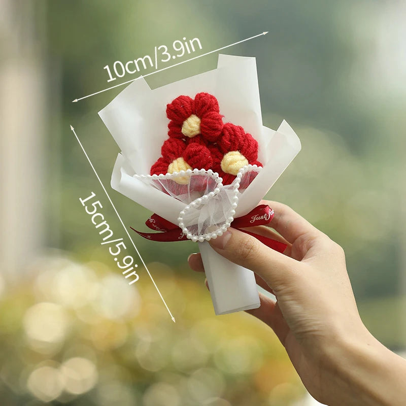 A hand holds a bouquet of handmade red and yellow crochet flowers wrapped in white paper and tied with a red ribbon.