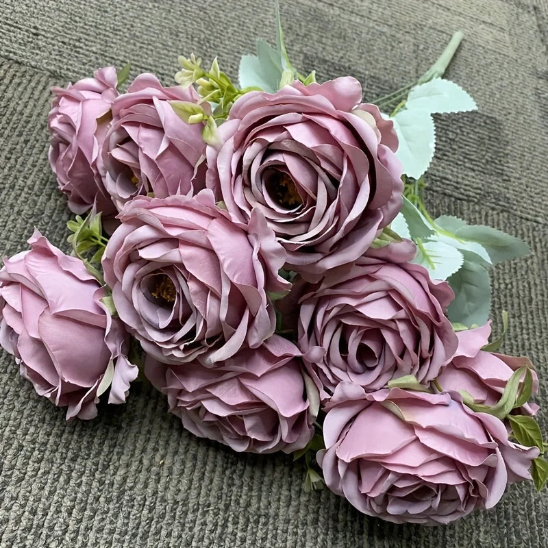 A bouquet of nine realistic silk roses in shades of dusty mauve and pink on a green fabric background.