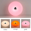 Drei Versionen einer donutförmigen, wiederaufladbaren Wandlampe in Weiß, Orange und Pink.