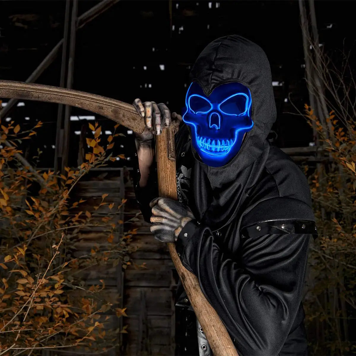 A person wearing a black hooded garment and holding a wooden branch, with a glowing blue skull mask on their face.