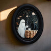 Oval framed wall art depicting a ghostly figure and a black cat in a dark floral pattern.