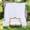 A white backdrop curtain decorated with greenery and pink flowers, featuring an antique wooden sofa and side tables outdoors.