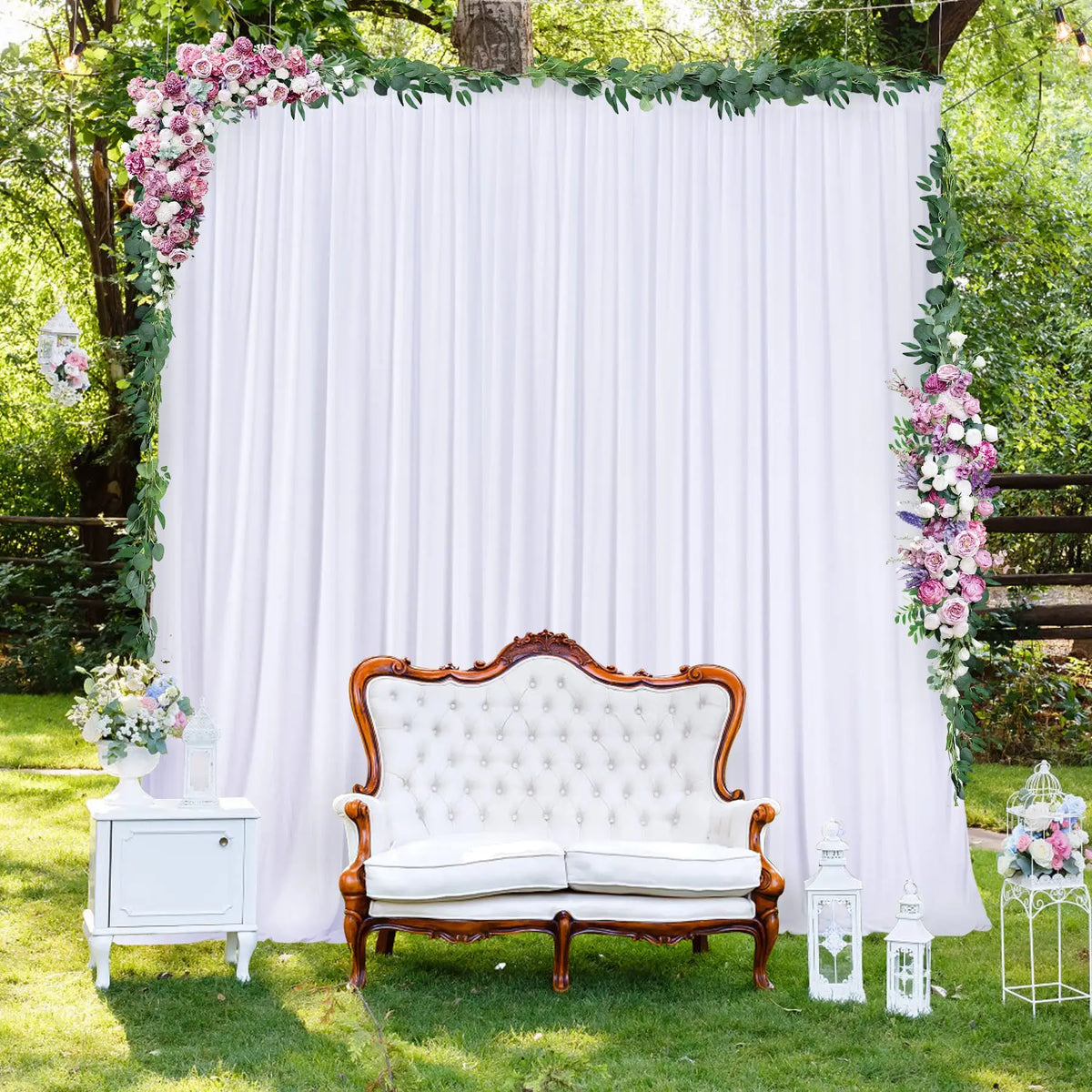 A white backdrop curtain decorated with greenery and pink flowers, featuring an antique wooden sofa and side tables outdoors.