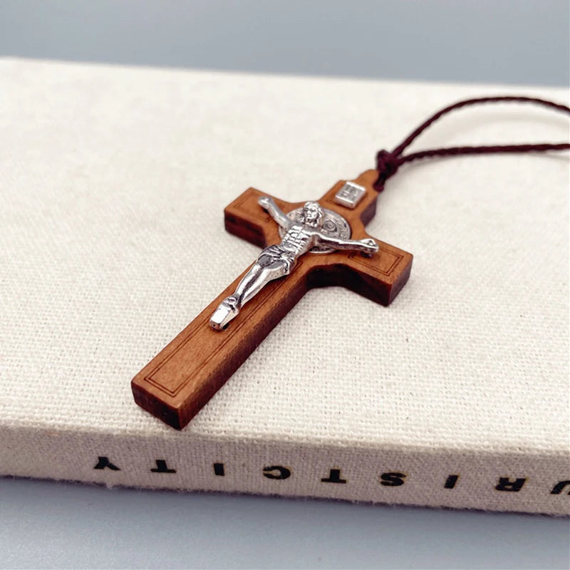 A wooden cross necklace featuring a figure of Christ, suspended by a dark brown waxed rope cord, resting on a light-colored f