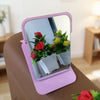 A pink foldable vanity mirror with a silver frame reflects a potted plant of red and yellow flowers.