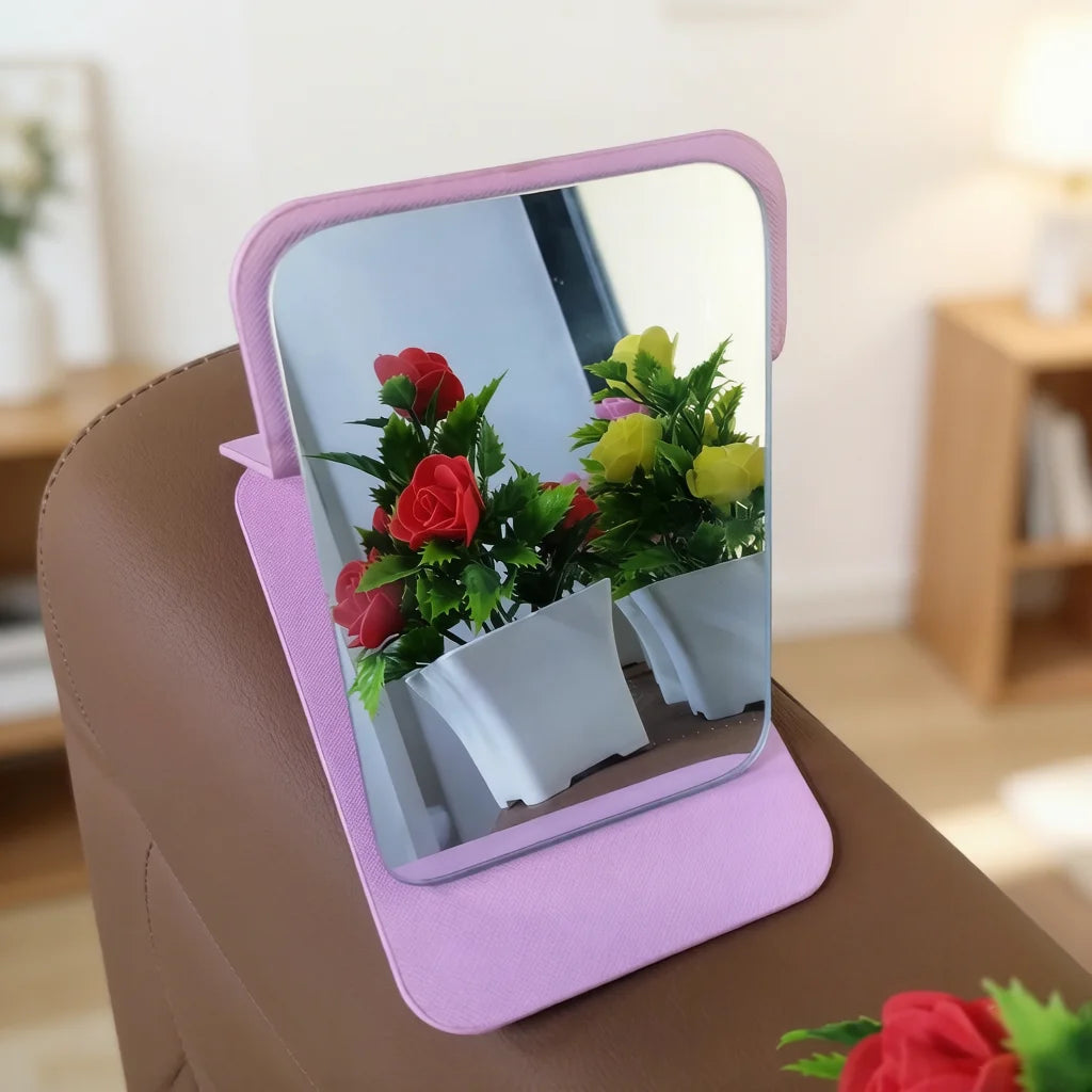 A pink foldable vanity mirror with a silver frame reflects a potted plant of red and yellow flowers.