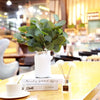 A white ceramic vase holds artificial eucalyptus stems on a wooden table.