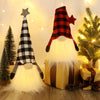 An illuminated LED gnome Christmas decoration with a plaid hat and white beard sitting on a yellow gift box.