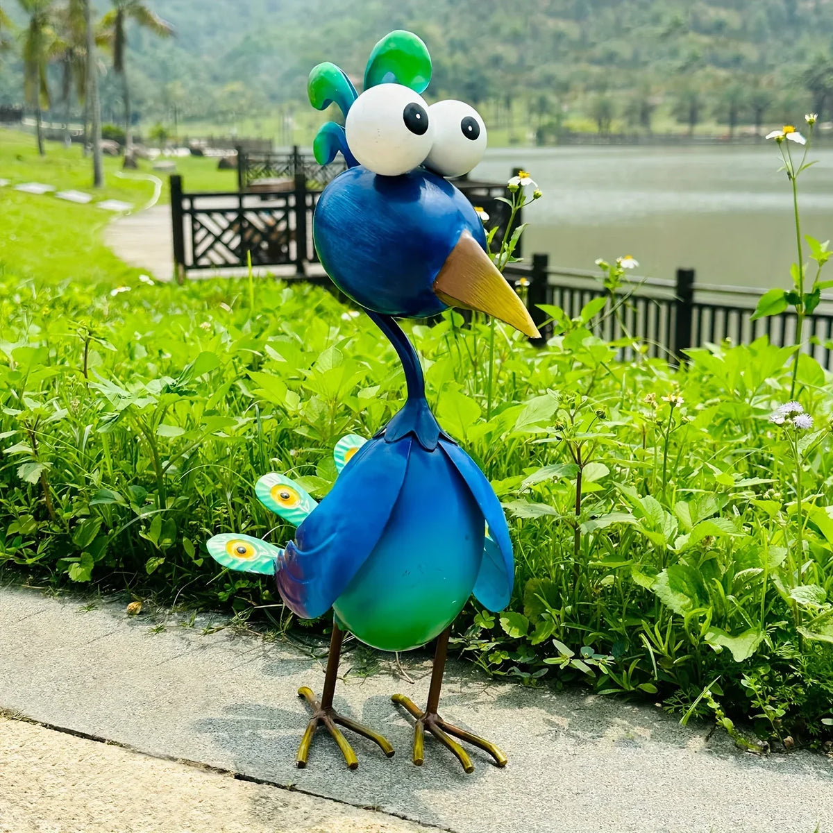 A vibrant blue metal peacock garden sculpture with large white eyes standing in green foliage near a body of water.