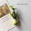 A small yellow ceramic vase holds a sprig of green artificial flowers on a white background.