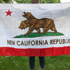 A white flag featuring a brown bear silhouette on a green hill and red star, with the text NEW CALIFORNIA REPUBLIC below.