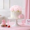 A white cake decorated with pink peonies sits on a stand next to small pink flowers and a pink birthday card.