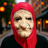 A woman wearing a red headscarf and a realistic full-face latex mask resembling an old, wrinkled face.