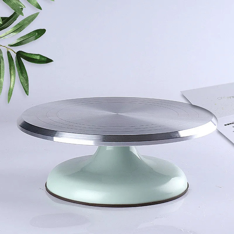 A silver aluminum cake turntable with a light mint green base is shown on a white surface.