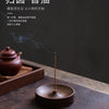 A dark brown solid wood incense stick burner with a thin metal stick emitting smoke on a wooden surface.